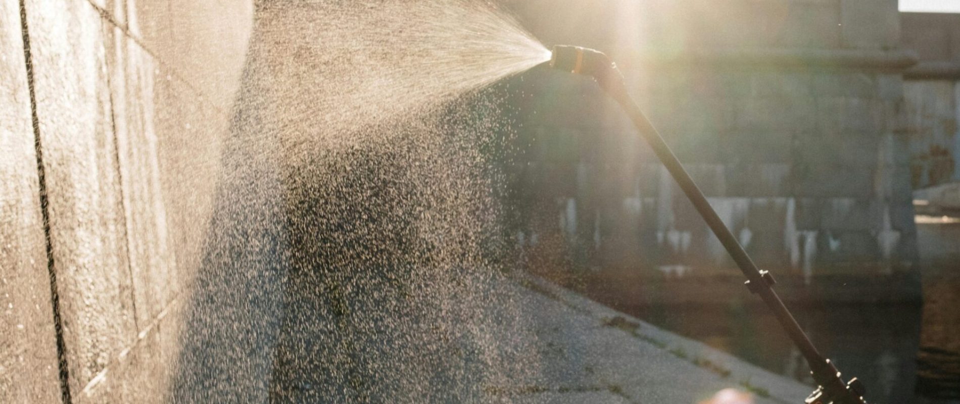 Man in orange uniform wearing facemask sprays water on wall outdoors, sunlight shining.